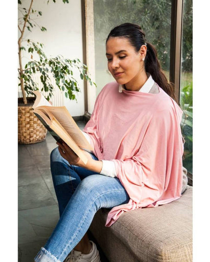 Mujer sentada en un sofá usando un pañuelo de lactancia color palo rosa de Amamantas mientras lee un libro.