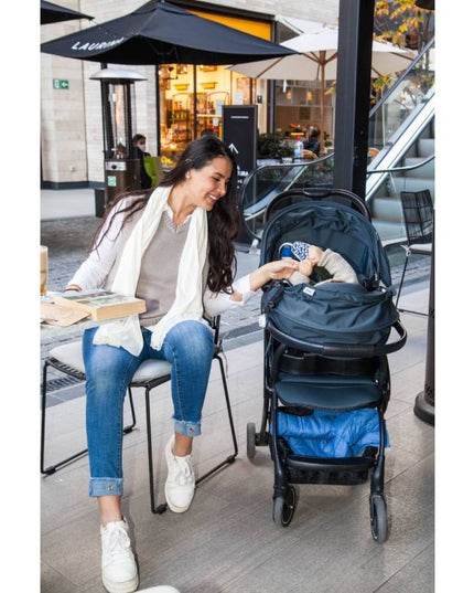 Madre usando un pañuelo de lactancia crudo de algodón en un café, cubriendo a su bebé en coche. Diseño versátil y elegante.