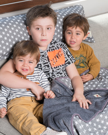 Tres niños sonrientes sentados en un sofá con un cojín XL de 90cm en color grafito con estrellas de Amamantas.